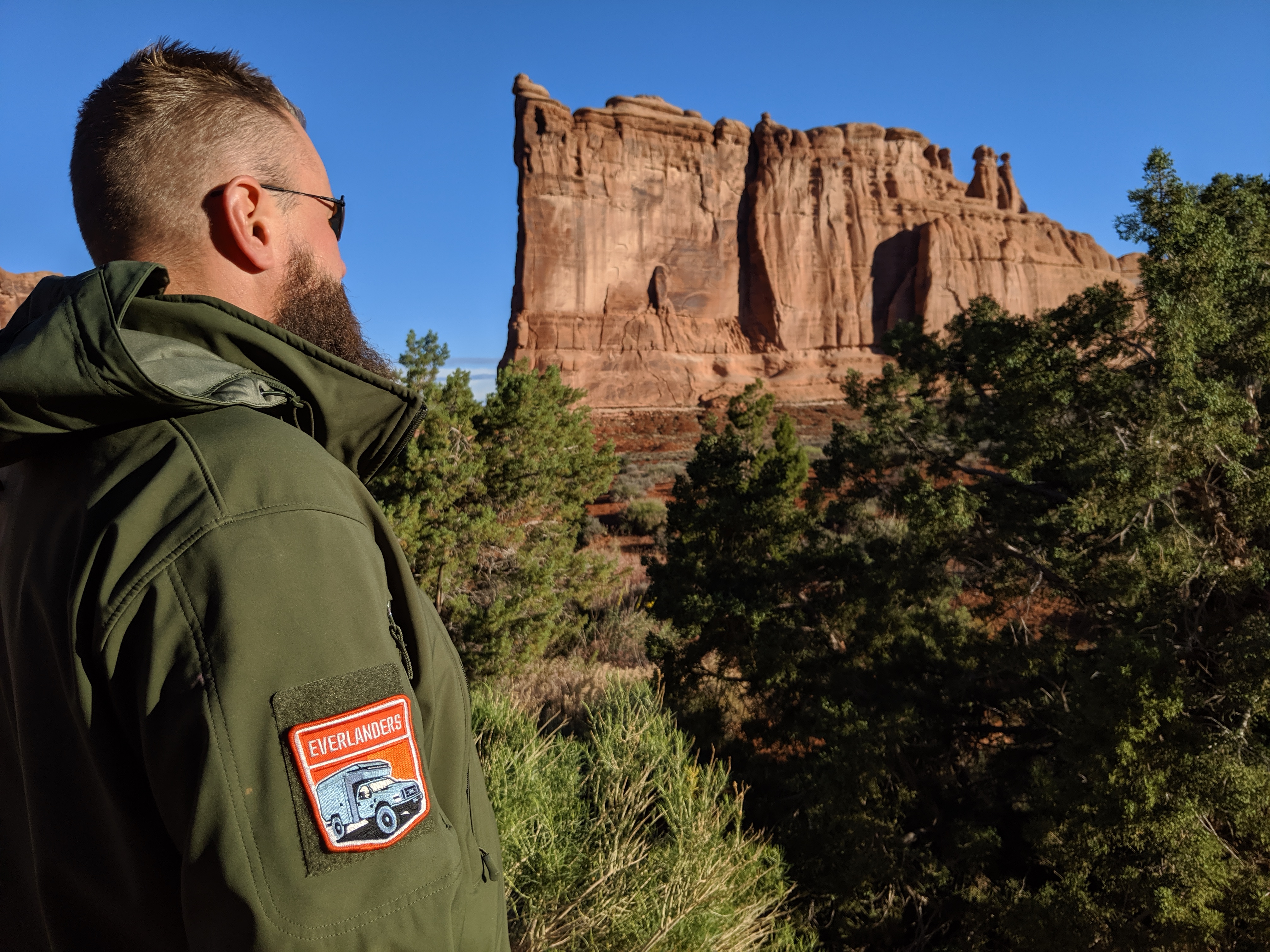 A person with short hair and a beard is standing in front of a large rock formation, wearing a green jacket with an 'Everlanders' patch, surrounded by trees under a clear blue sky.