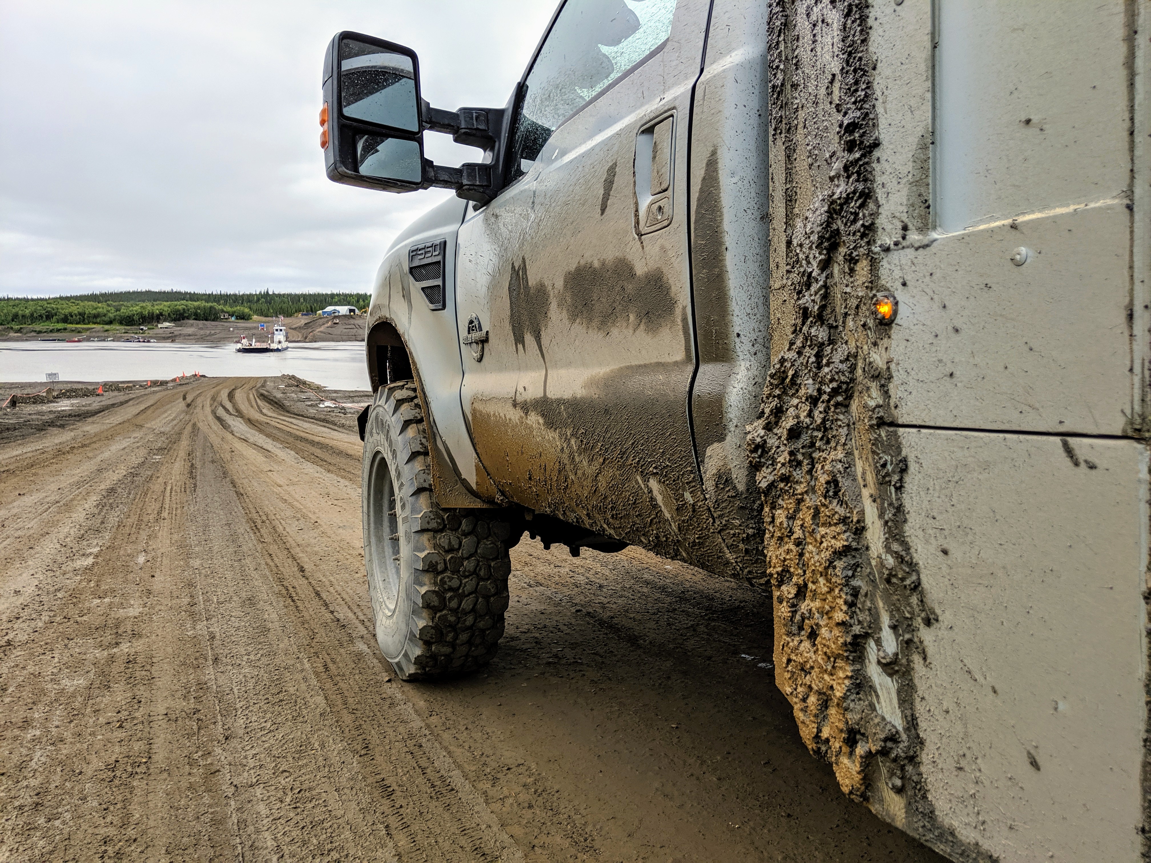 Close-up of a muddy truck parked on a dirt road with a view of a calm body of water and forested land in the background.