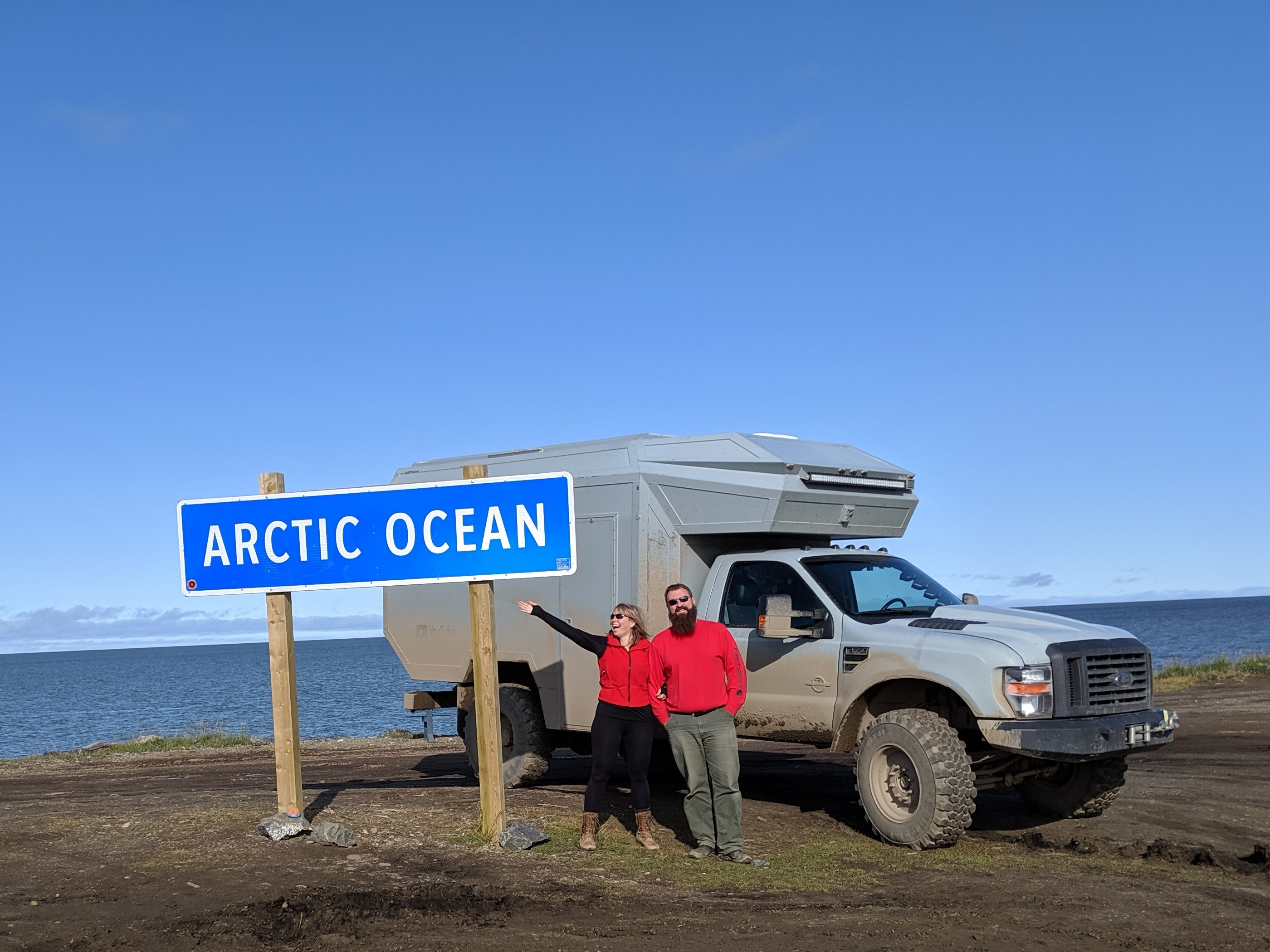 A woman and a man stand next to a rugged truck, pointing at an 'ARCTIC OCEAN' sign beside a body of water on a clear day.