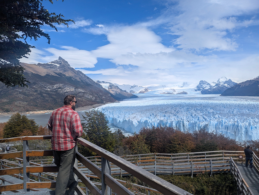Jason standing on a wooden walkway overlooking a massive glacier under a blue sky with scattered clouds and mountains in the background.