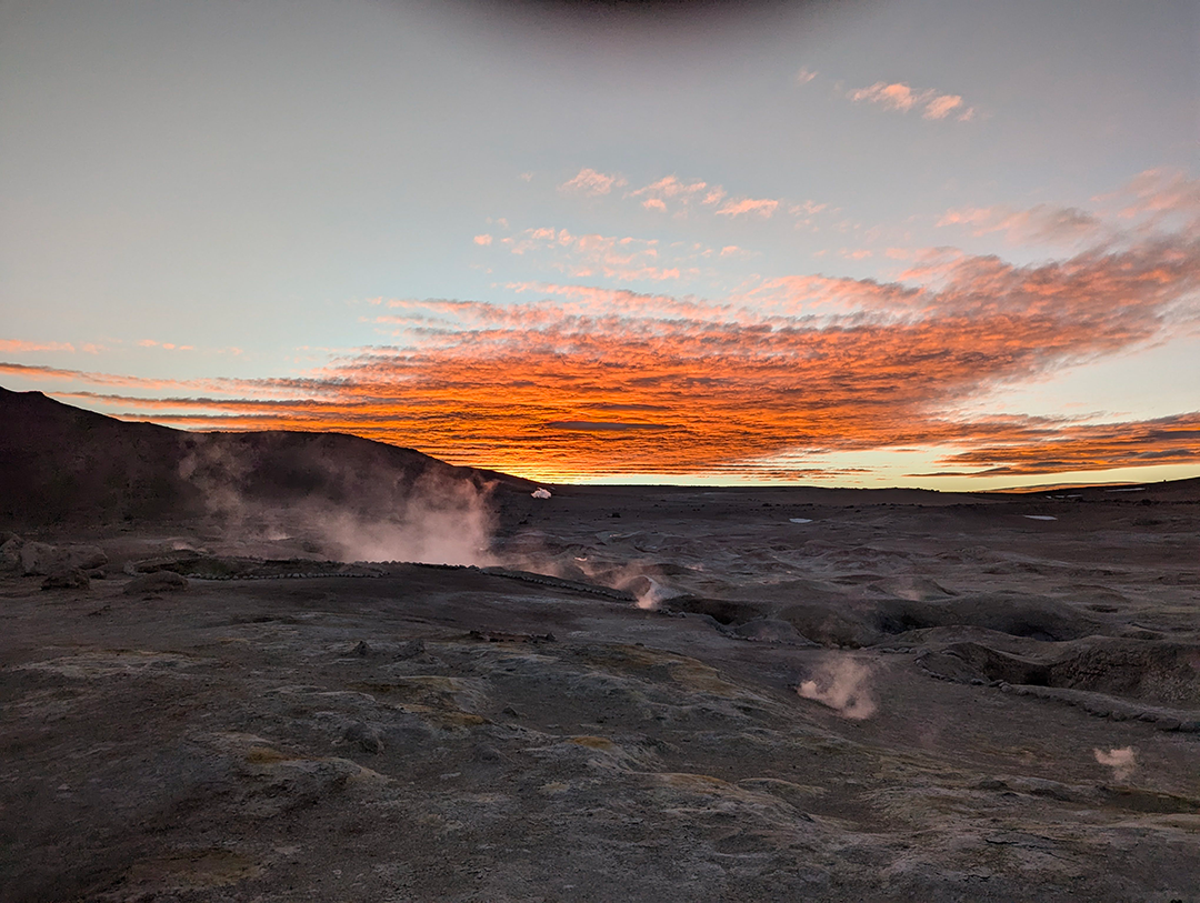 A stunning sunset over a mountainous landscape, with steam rising from geothermal vents creating a surreal atmosphere.