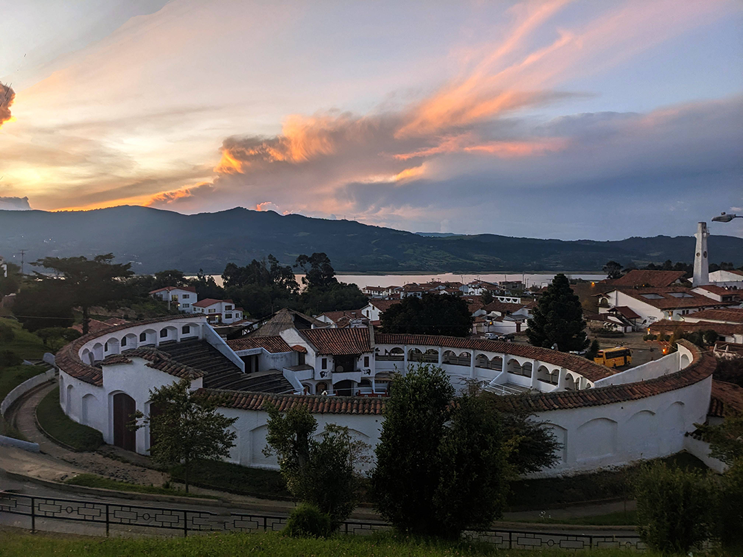 A panoramic view of a circular building set against a sunset backdrop, surrounded by lush greenery and mountains.