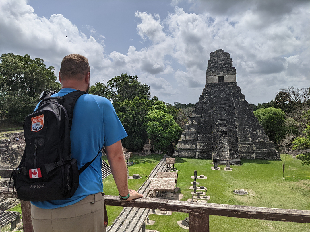 Jason standing in front of the Tikal temple in Guatemala, looking at the ancient structure surrounded by lush greenery under a cloudy sky.