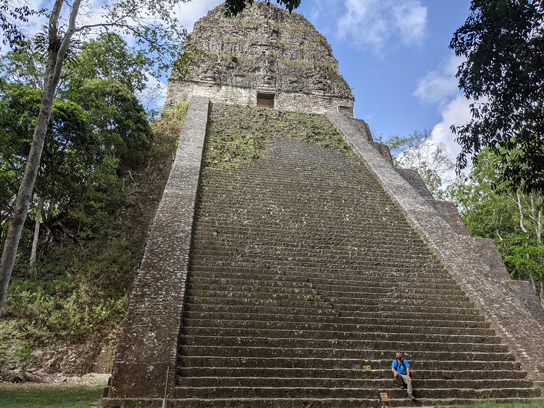 A large, ancient Tikal pyramid surrounded by lush vegetation, with a Jason sitting at the base of the steps.