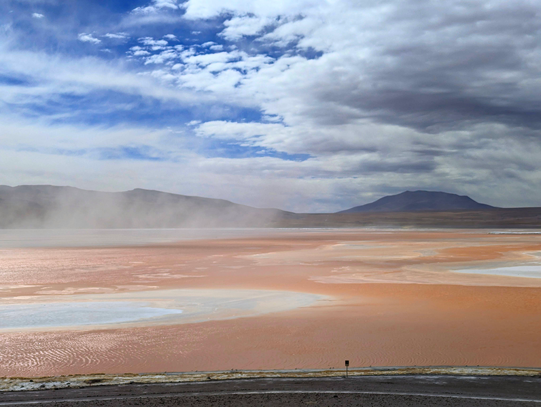 A stunning landscape featuring a pink-orange lake surrounded by mountains under a cloudy sky, evoking the surreal beauty of Bolivia's natural scenery.