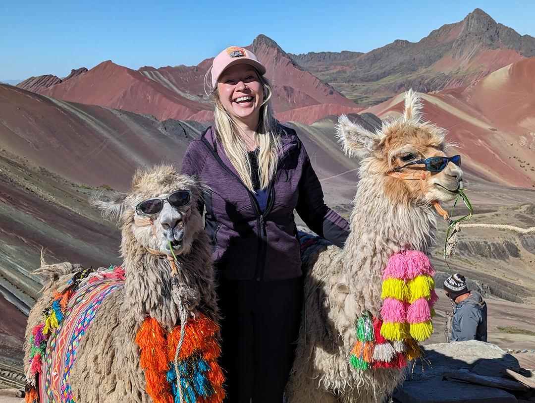 Kara smiling and posing with two llamas wearing sunglasses and colorful accessories, set against a backdrop of the vibrant mountain landscape.